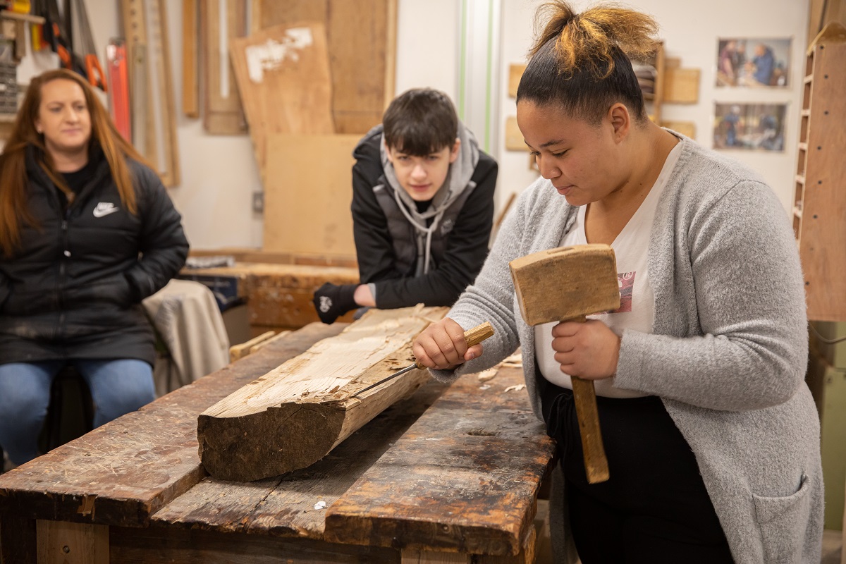 Two young people in our workshop. One is sat down and watching as the other uses a mallet and chisel to chip away at some wood.