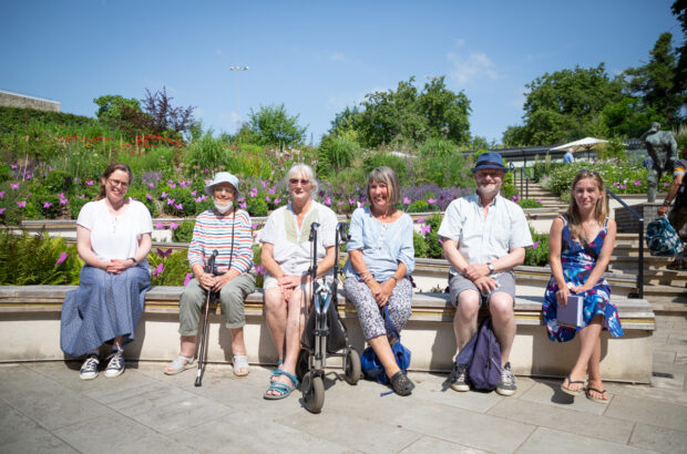 6 people sat on the rim of a wall in the walled garden at the Museum in the Park, Stratford Park.