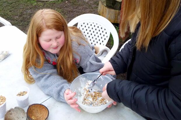 Two girls are at a table, one holding a bowl as the other uses two spoons to mix fat and seed together.