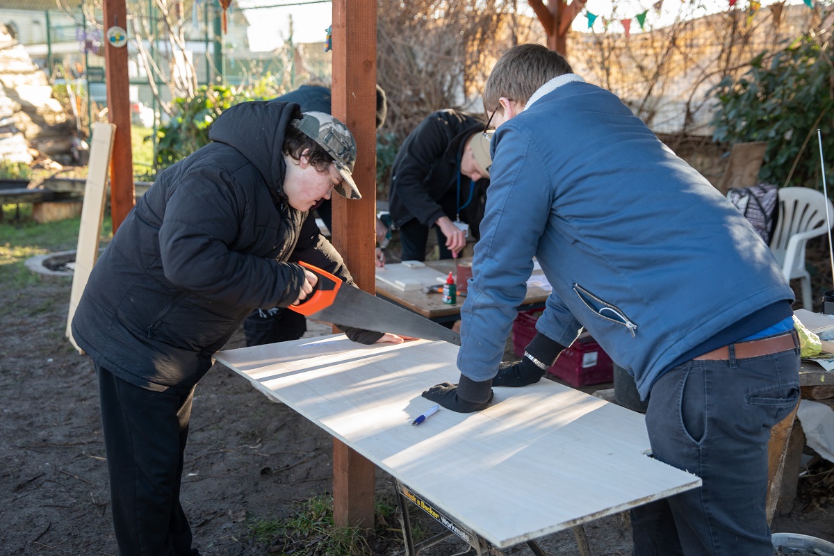 Two young people are our allotment. One is holding down a sheet of wood, the other is using a saw to cut it.