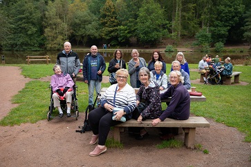 Group photo of everyone on a trip. Some are standing, some are sitting on benches. There are trees and a lake in the background.