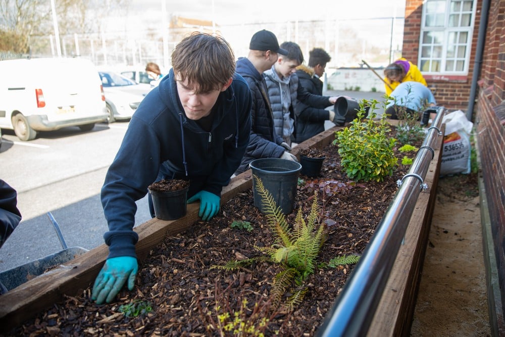A row of five young people alongside a wooden planter, shifting the soil and planting various plants.