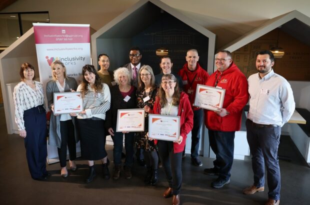 A line up of people smiling inside Gloucester Services and holding framed certificates showing their Inclusivity Works certificate.