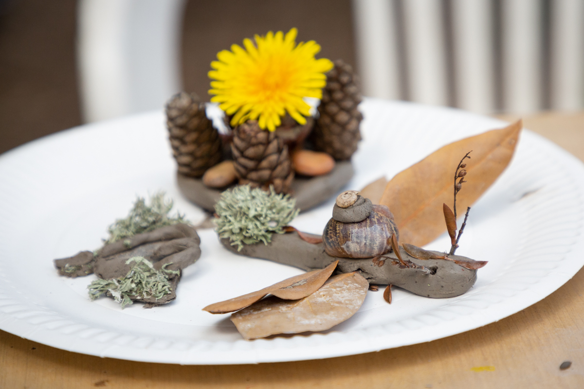 A close up of a paper plate with three clay statuettes on. At the forefront is a dragonfly which has used leaves for wings.