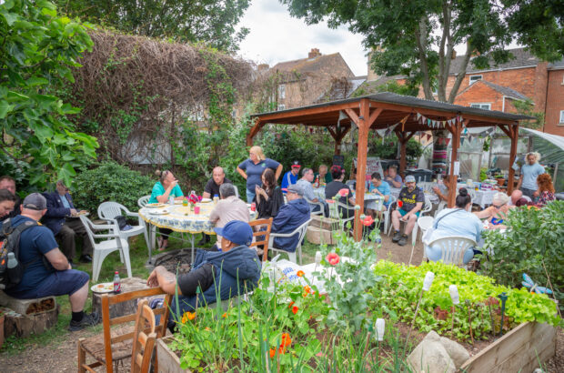 A wide shot of our alloment full of people. Some are sitting down at tables eating and chatting, others are standing up.