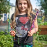 A young girl smiling at the camera as she holds 2 sticks with wool and ribbon wrapped around them.