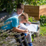 Two young boys sat on a bench in the allotment, writing on the 'Summer Trail' activity sheet using a pencil.