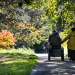 Two people, one in a mobility scooter, walking down a shaded path surrounded by trees with their backs to the camera.