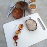 A table with two cups of seeds, some spoons, two coconut halfs, and a chopping board on it. On the chopping board is half a coconut filled with fat and seeds as well as a needle and thread with dried fruits and popcorn on it.