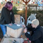 One young person holding some roofing on top of a wooden box as another person hammers nails in to keep it in place.