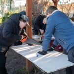 Two young people are our allotment. One is holding down a sheet of wood, the other is using a saw to cut it.