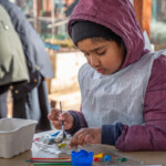 Child in a plum-coloured coat sat at a table painting his pebble yellow.