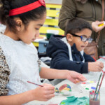 Two children sat at a table painting pebbles. The girl in the forefront is on her third pebble.