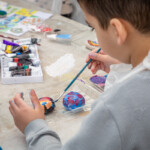 Over the shoulder shot of a child using a brush to paint a pebble. There's lots of paint on the table.