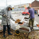 Two men with shovels are shovelling soil from bags into a wheelbarrow, in the Friendship Cafe parking lot.