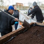 Three young people drag a bag full of soil toward a planter. One young man in the forefront with a space is getting ready to shovel the soil.