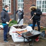 Three young people around a bag of soil, using shovels to move the soil into a wheelbarrow.