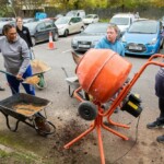 Five people standing in the Friendship Cafe parking lot. One lady is using a shovel to move gravel from a wheelbarrow into an orange mixer.