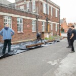 Photo showing the side of the Friendship Cafe building, in their car park. 5 People are standing there and working on building some planters,