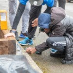 A young person crouched down using a drill to screw two planks of wood together.
