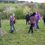 Group of young people in a field. They are watching a demonstration being given by an older lady.