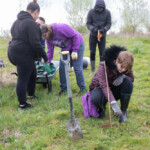 Group of 5 people in a field. The person in the front is crouched down and placing soil in a hole where a tree has just been planted.