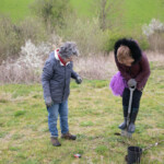 Two women in a green field which is Gloucester Services Growing Space. The older woman is watching a younger woman use a shovel to dig a hole.