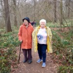 Two older ladies smiling at the camera as they walk along a path in a forested area.