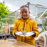 Young boy in a yellow coat smiling at the camera and holding a paper plate with his clay statuette on.