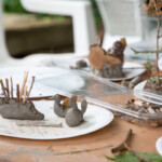 A messy table with some clay statuettes. A clay hedgehog with sticks as spines, a bunny, a duck with leaves for wings.