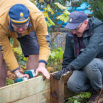 Two Spirax Sarco volunteers fixing the wooden border of a plant bed. One is holding the wood in place while the other drills it.