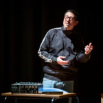 JPDL standing behind a desk with audio equipment on it, in the middle of his "Here He Stood" performance.