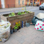 The finished planter with a bag full of weeds next to it, as well as some watering cans and a wheelbarrow.