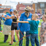 A young boy holding back the string of his bow, with a young girl and the archery instructor observing him.