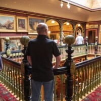 Gary and Pete, pictured from behind, looking over the bannister inside a manor house with paintings on every wall.