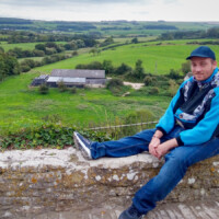 A participant smiling at the camera as he sits on a stone wall overlooking rolling green hills.