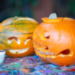 Two pumpkins on a table. The one in the foreground has a wriggly mouth and crescent moon eyes, the one in the background has a smile and curved eyes.