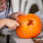 A girl carving out an 'x' shaped eye on a pumpkin.