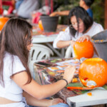 Two girls sat at a table, the one in the foreground is drawing a design on her pumpkin while the one in the background is carving.
