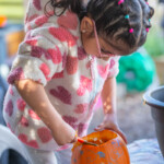 A young girl stood up and scooping out the insides of a pumpkin with drawings on it.