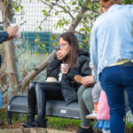 A young girl sitting on a bench in the allotment, eating a breadroll and holding a cup of soup.