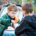 Two kids eating soup with bread rolls, one has his back to the camera. The one facing the camera is using his spoon to scoop up some soup.