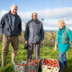 Michael, Chrissy, and Mr.Patel smiling at the camera as they stand infront of the apple orchard and behind some crates of apples.
