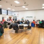 A wide shot of the hall, showing all the participants sat down at the tables.