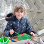 A child smiling at the camera as she paints her paper plate.