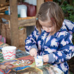 A young girl scooping up some glue from a pot so she can stick beans to her template.