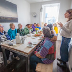 Everyone sat around a table eating croissants and drinking some tea. They're listening to Clare talk about the schedule for the day.