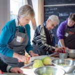 Various people around the table working on their assigned recipe. One woman is chopping up a cabbage,