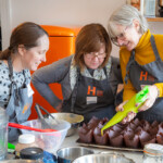 Three people making cupcakes. One in a yellow jumper is piping the mixture into the brown cake cases while the other two watch.