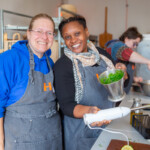 Two group members in charge of the pesto. They're smiling at the camera, with the one on the right holding up the metal jug which they've been blending the herbs in.